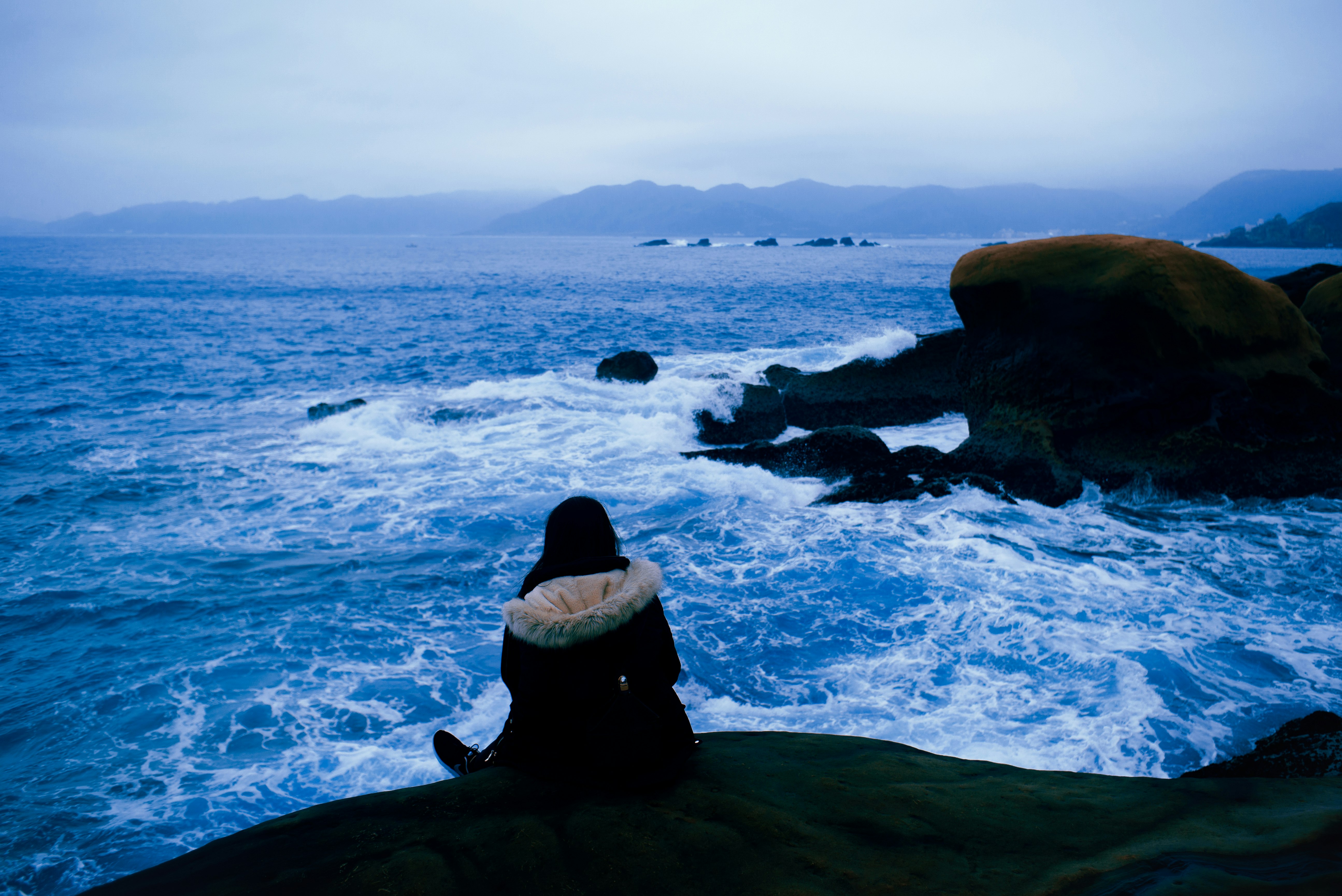 man in white and black jacket sitting on rock formation near sea during daytime